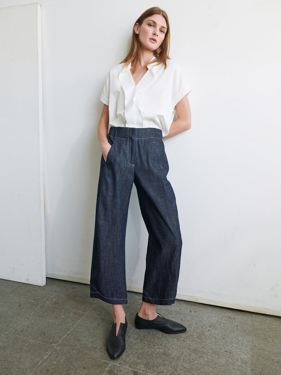 A person with shoulder-length brown hair stands in a relaxed posture against a white wall. They are wearing a white short-sleeved shirt tucked into high-waisted, wide-leg Aissa Cargo Pants by Zero + Maria Cornejo and black slip-on shoes. Their hands are casually in the pockets of their pants.