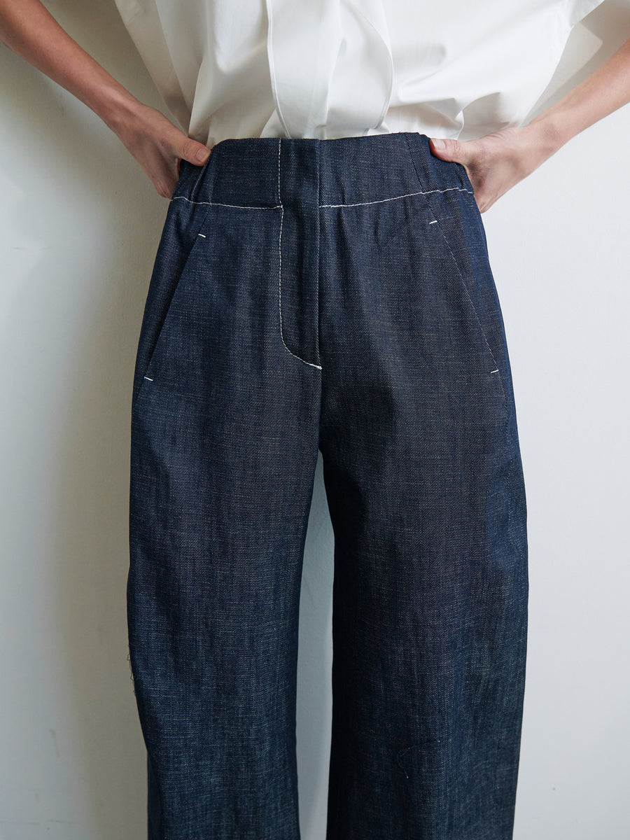 A person wearing a white shirt and high-waisted, loose-fitting Aissa Cargo Pants by Zero + Maria Cornejo is shown from the torso down. The hands are positioned on their hips, emphasizing the wide-leg style of the pants. The backdrop is plain and light-colored, highlighting the elegance of these GOTS-certified cotton trousers.