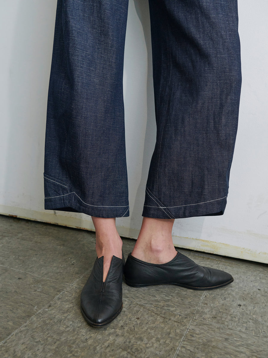 A person wearing dark blue, cropped wide-leg Aissa Cargo Pants by Zero + Maria Cornejo stands against a plain wall. They are also wearing black slip-on shoes with an unusual v-shaped cut-out near the top. The flooring is gray and slightly textured. Only the person's lower legs and feet are visible.