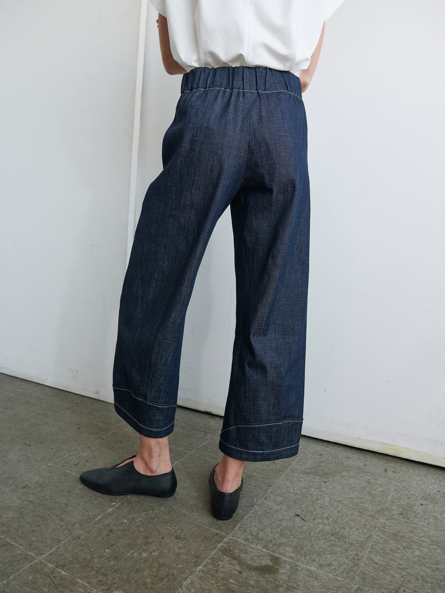 A person wearing the Aissa Cargo Pant by Zero + Maria Cornejo, made of navy blue GOTS-certified organic cotton denim with an elastic waistband, and black shoes stands on a gray floor with a white wall background. Their back is facing the camera, and they are also wearing a white top. The pants reach their ankles.