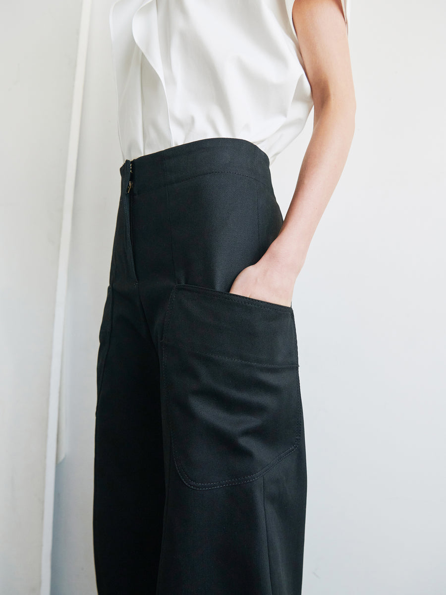 A person wearing a white sleeveless top and black Curved Eda Culotte high-waisted organic denim pants from Zero + Maria Cornejo, which feature large pockets, has their hand in one of the pockets. The image focuses on the torso and partial arm, highlighting the clothing details.