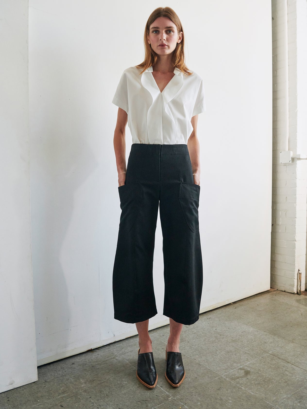 A person with long hair is standing indoors against a white wall and door. They are wearing a white short-sleeve button-down shirt tucked into the Curved Eda Culotte by Zero + Maria Cornejo, high-waisted and wide-leg organic denim pants paired with black heeled mules. The overall look, featuring GOTS-certified cotton pieces, is casual yet stylish.