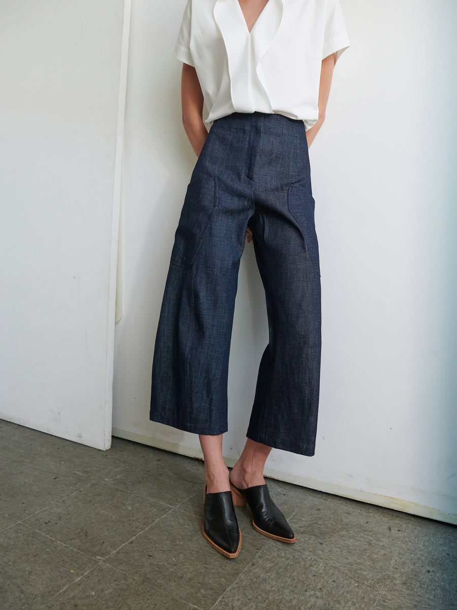 A person is standing against a white wall, wearing a white short-sleeve blouse tucked into the Curved Eda Culotte by Zero + Maria Cornejo, which are high-waisted and made of GOTS-certified organic denim in dark blue with wide-leg cropped hems. They are also wearing black mules. The floor is tiled in a neutral color. The person's head is not visible in the frame.