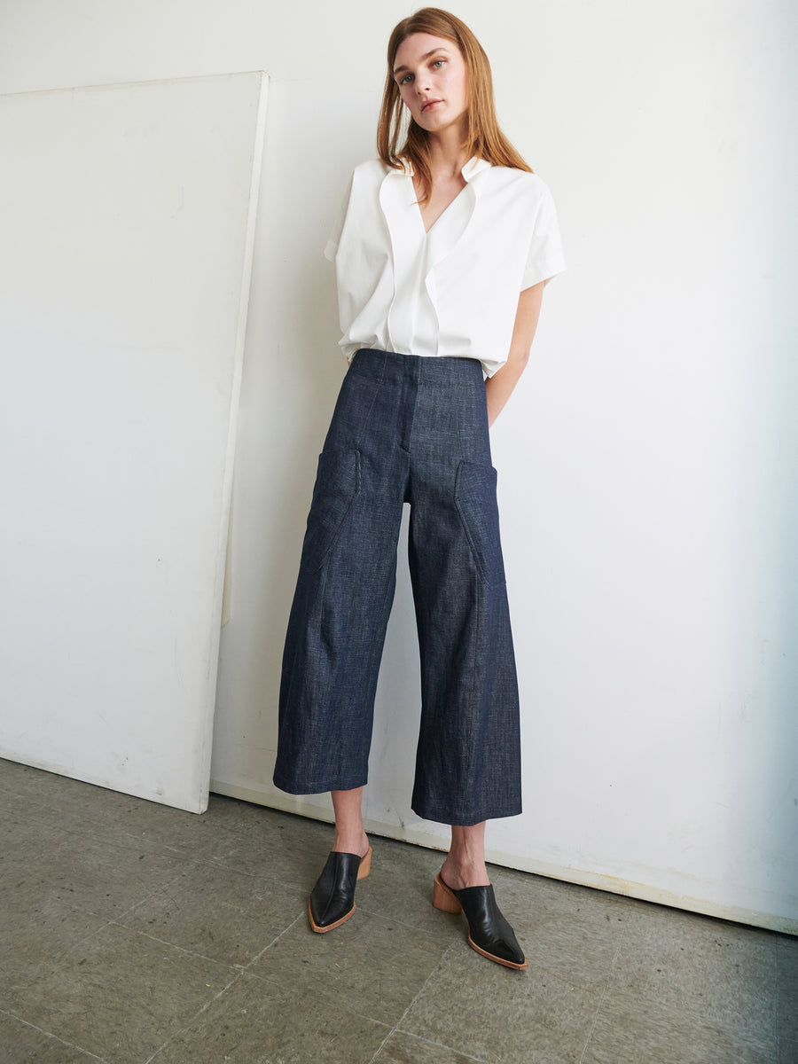 A person with long hair is standing indoors against a white wall and door. They are wearing a white short-sleeve button-down shirt tucked into the Curved Eda Culotte by Zero + Maria Cornejo, high-waisted and wide-leg organic denim pants paired with black heeled mules. The overall look, featuring GOTS-certified cotton pieces, is casual yet stylish.