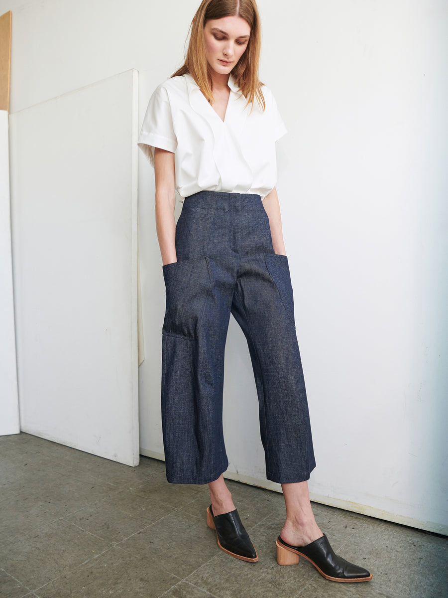 A woman with straight blonde hair stands indoors against a white wall. She is wearing a white short-sleeved blouse tucked into high-waisted Curved Eda Culottes from Zero + Maria Cornejo, featuring large pockets and cropped wide legs. She also wears black mules with a wooden heel.