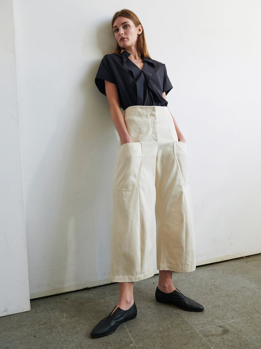 A woman stands against a white wall, wearing a loose black short-sleeve shirt tucked into Zero + Maria Cornejo’s Curved Eda Culotte—organic high-waisted cream denim with wide legs and large front pockets—and black flats, looking relaxed with hands in her pockets.