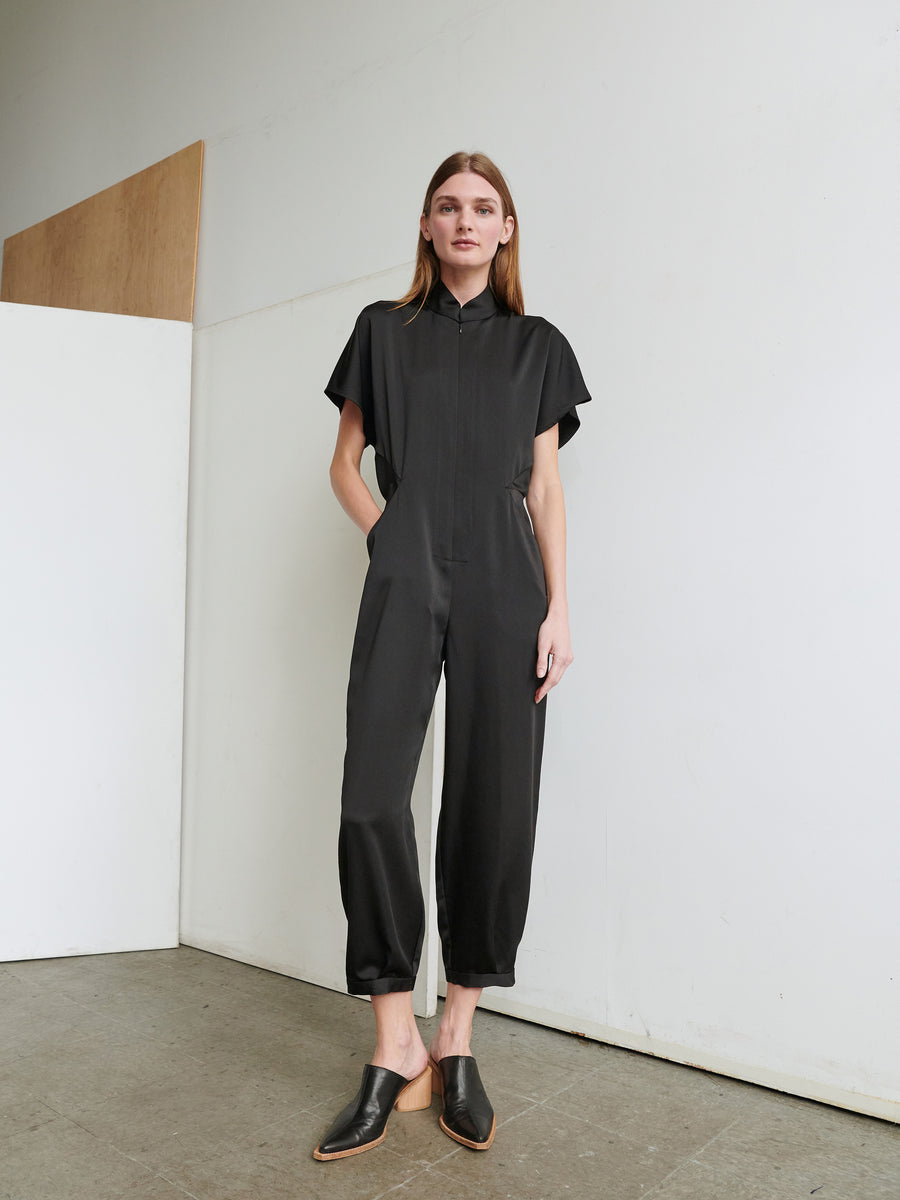 A woman stands indoors against a white wall, dressed in the black Elia Jumpsuit by Zero + Maria Cornejo, featuring short sleeves and ankle-length pants. Her hands are in her pockets, and she is wearing black slip-on shoes. She has long, straight hair and a neutral expression.