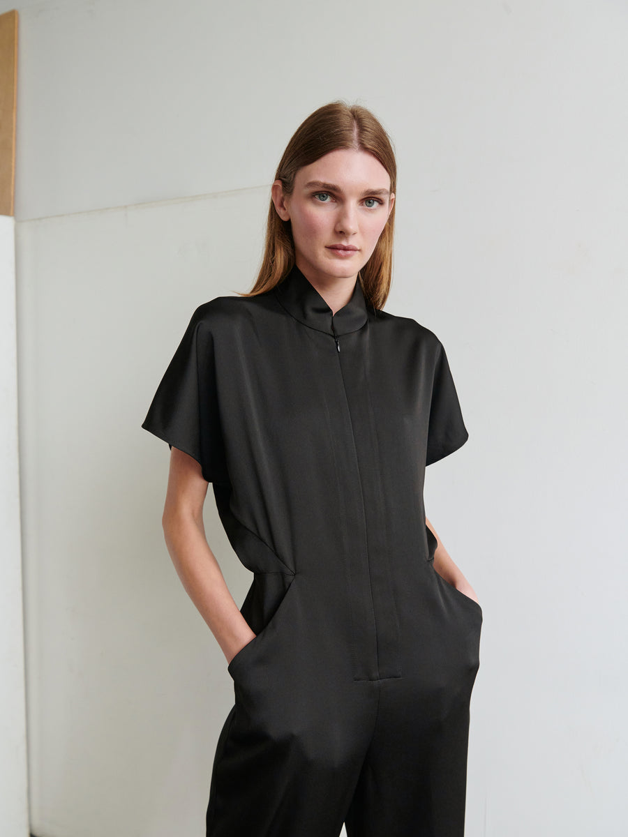 A person with long hair is wearing the Elia Jumpsuit by Zero + Maria Cornejo, a sleek, black recycled drape jumpsuit made of 100% recycled fabric, hands in pockets. The background is minimal and white with a wooden detail on the left. The overall look is stylish and modern, reflecting its made-in-New York origins.