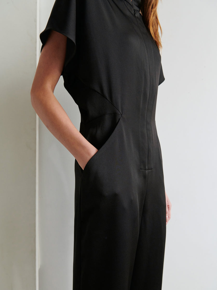 A person is wearing the sleek, black Elia Jumpsuit from Zero + Maria Cornejo, with their left hand in their pocket. The image focuses on the upper torso and upper legs, showcasing the elegant, minimalist design of this recycled drape jumpsuit made in New York. The background is plain and light-colored.