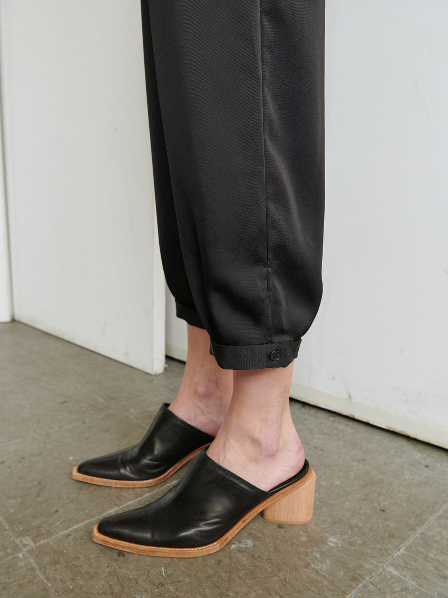 A person dressed in the Elia Jumpsuit by Zero + Maria Cornejo, which features black, cuffed, ankle-length pants made from 100% recycled fabric, and wearing black heeled slip-on shoes with a wooden block heel and pointed toes stands on a gray floor. The background includes a white wall and a doorframe.