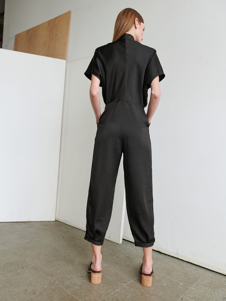 A person with long hair, shown from the back, stands in a minimalistic room. They are wearing the black Elia Jumpsuit with cropped legs from Zero + Maria Cornejo and wooden platform sandals. The room features light-colored walls and a concrete floor, creating a serene atmosphere.