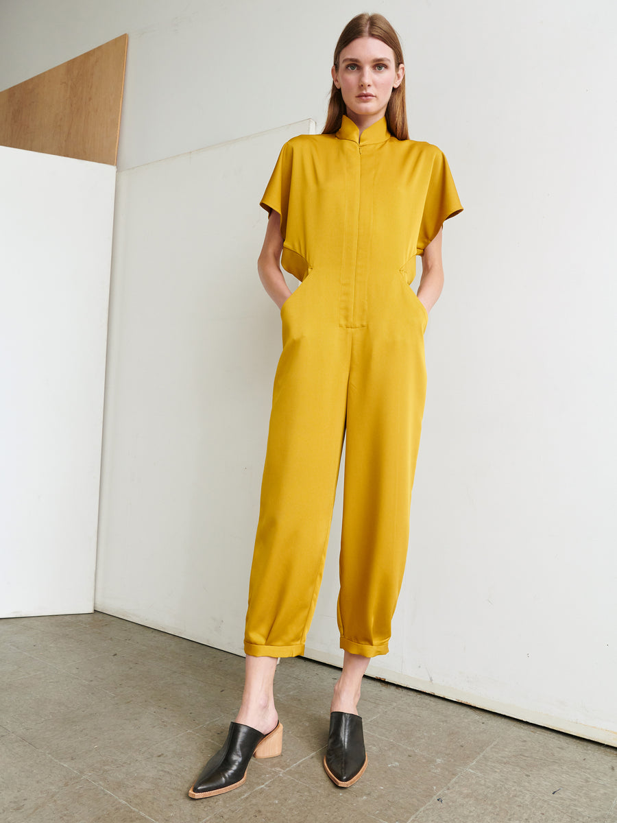 A woman with long hair stands against a white wall, wearing the mustard-colored Elia Jumpsuit with short sleeves and cropped legs from Zero + Maria Cornejo. She has her hands in the pockets and is wearing black slip-on mules with a low heel. The floor is a neutral-colored tile.