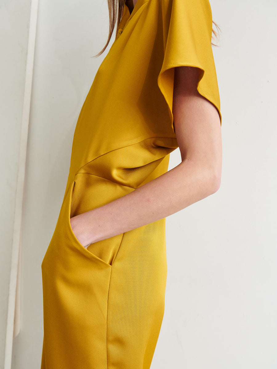 A person wearing the Elia Jumpsuit in mustard yellow, made by Zero + Maria Cornejo from 100% recycled fabric, with one hand in the side pocket. The image shows a close-up of the side and part of the arm, focusing on the jumpsuit's sleeve and pocket detail. The background is a plain, light-colored wall.