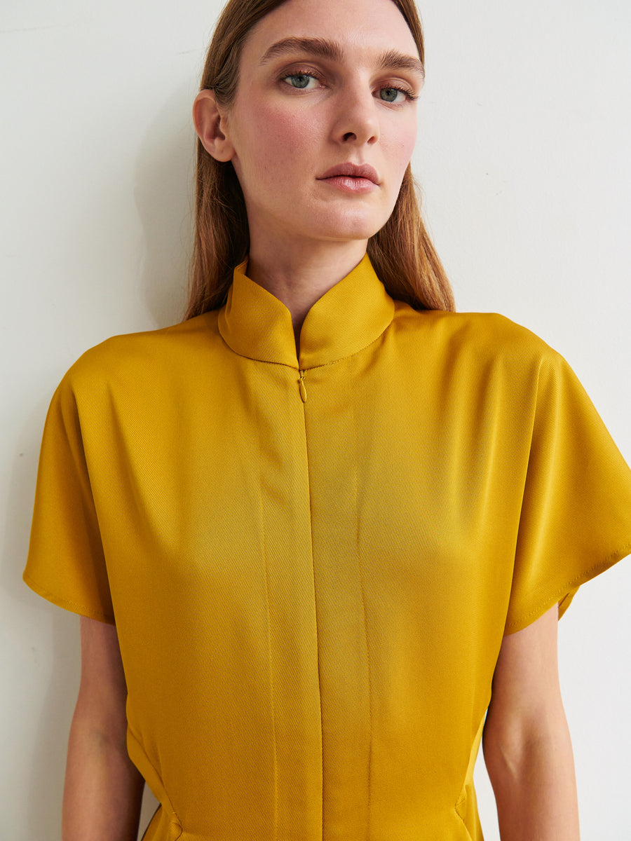 A person with long brown hair stands against a plain background, wearing the Elia Jumpsuit by Zero + Maria Cornejo, which features a mustard yellow color, short sleeves, and a high neck made from 100% recycled fabric. The person looks directly at the camera with a neutral facial expression.