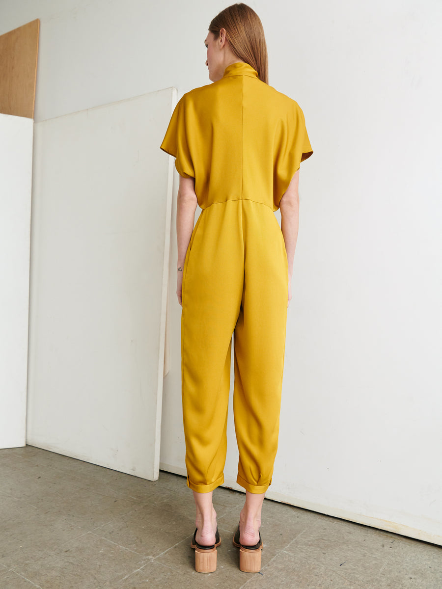 A woman stands facing a white wall, wearing the Elia Jumpsuit from Zero + Maria Cornejo. The jumpsuit is mustard yellow, made from recycled materials, and features short sleeves and loose-fitting legs. She has light brown hair and is also wearing platform sandals. The setting appears to be a minimalistic indoor space with a concrete floor.