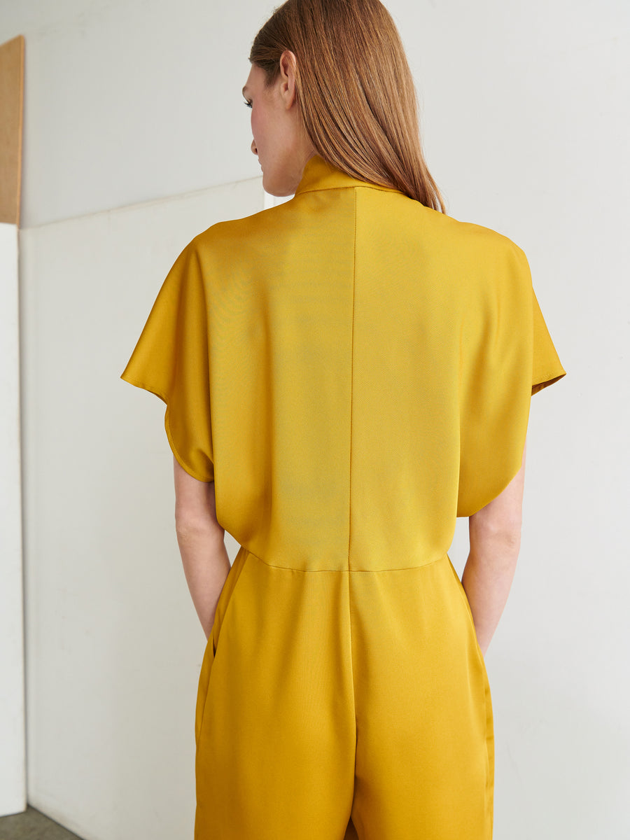 A person with long, straight hair is standing with their back to the camera, clad in a mustard yellow, short-sleeved Elia Jumpsuit by Zero + Maria Cornejo. The background is a plain, light-colored wall.