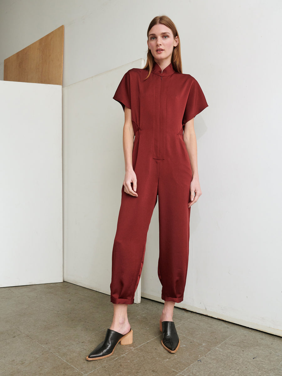 A person wearing the Elia Jumpsuit by Zero + Maria Cornejo, a maroon-colored ensemble made from recycled materials, stands against a minimalist backdrop featuring white and wooden panels. The jumpsuit, crafted in New York, showcases short sleeves and a collar. The person exudes a relaxed demeanor with hands tucked into pockets and eyes gazing slightly to the side while donning black mules.