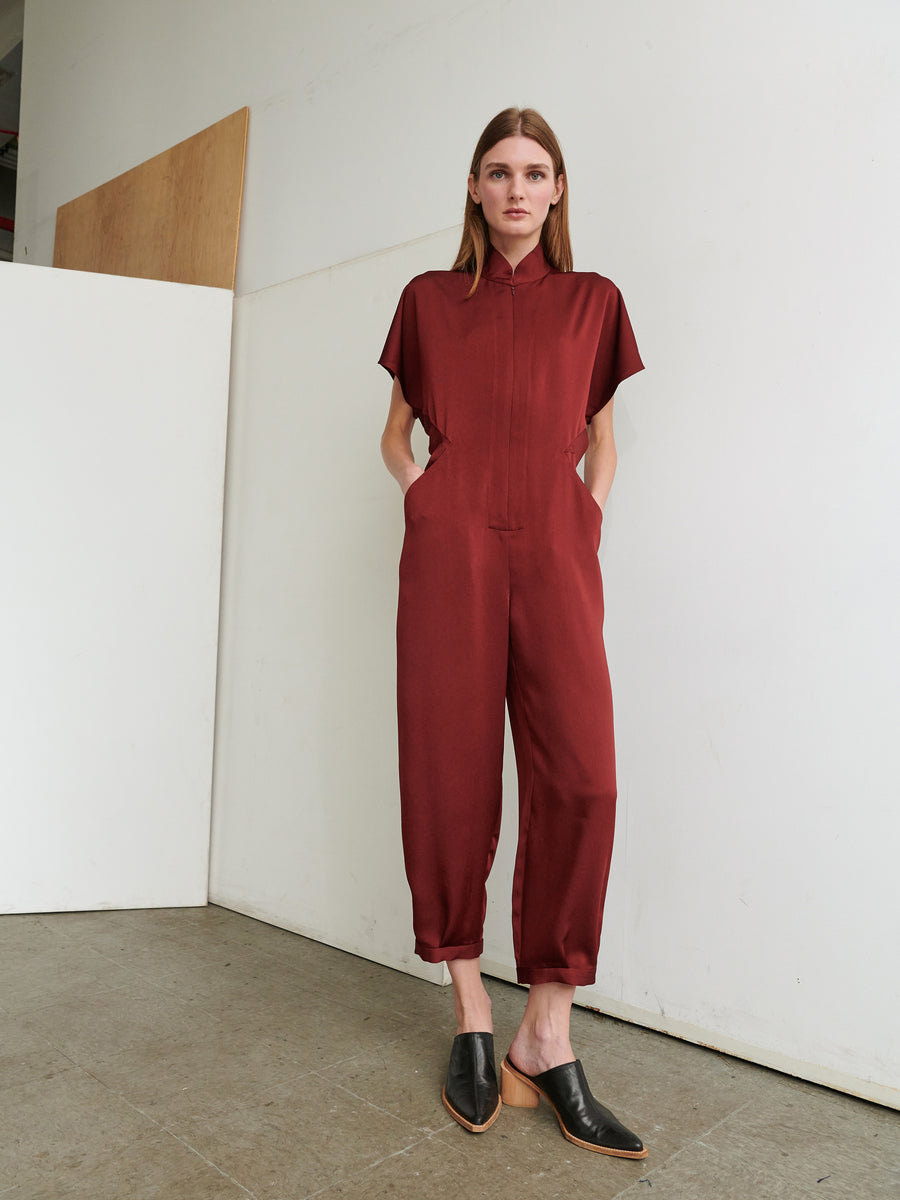 A woman stands indoors against a plain white wall and wooden panel, wearing the Elia Jumpsuit by Zero + Maria Cornejo in maroon, paired with black slip-on shoes. She has her hands in the jumpsuit pockets and looks straight ahead. The floor is made of gray concrete tiles.