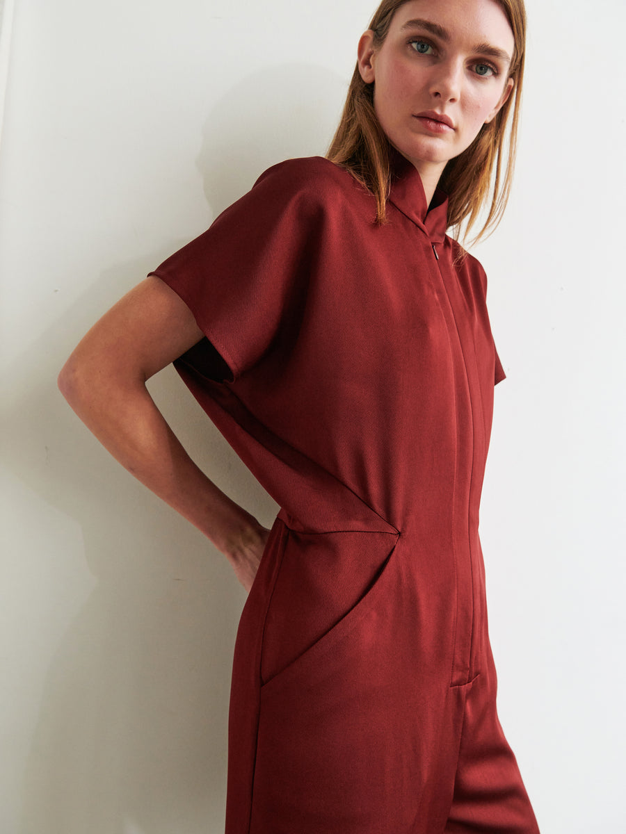 A person stands facing the camera, dressed in the short-sleeved, 100% recycled fabric Elia Jumpsuit by Zero + Maria Cornejo, with hands tucked in front pockets. They have light brown hair and are positioned against a neutral background, gazing directly at the camera.