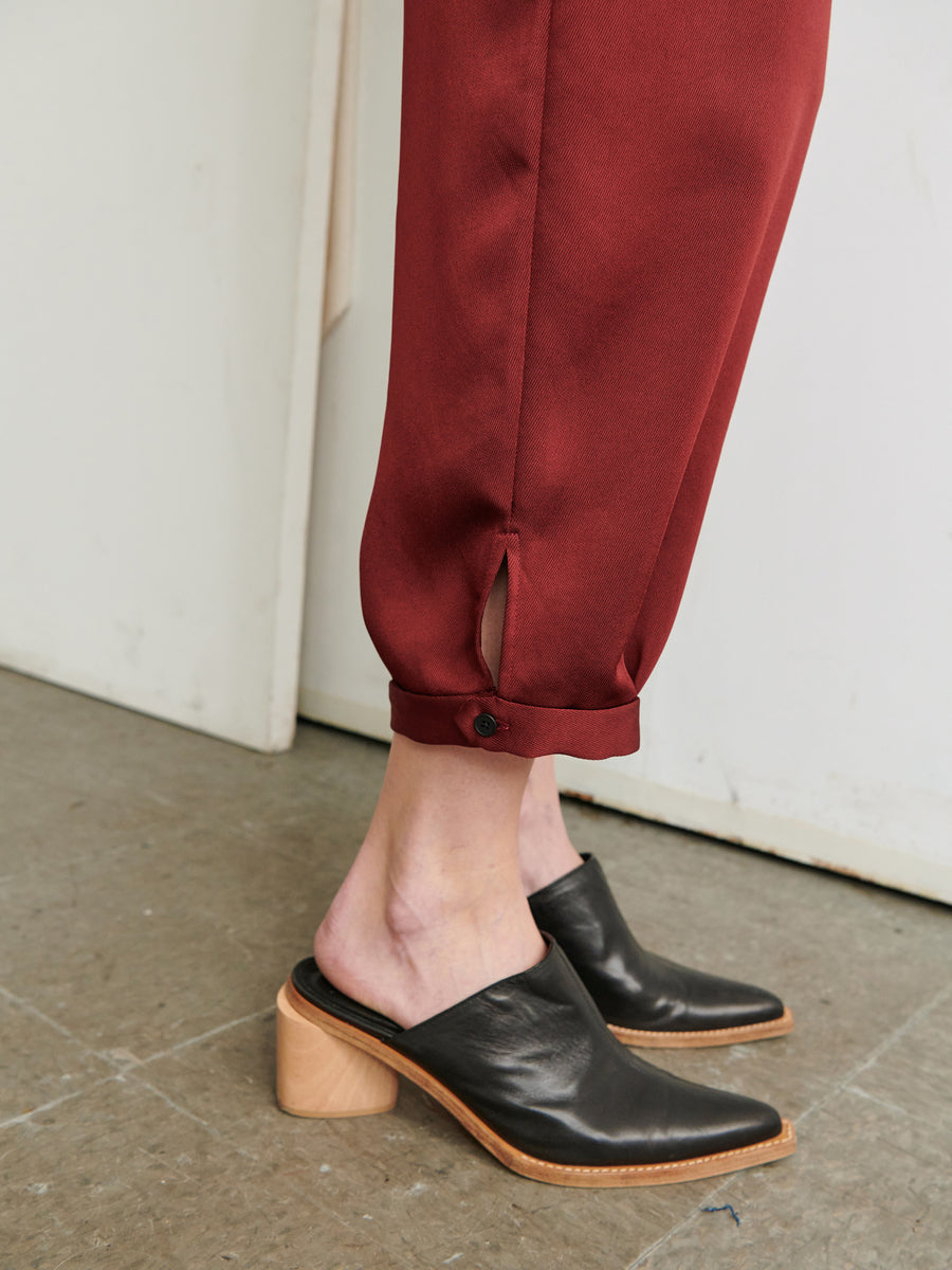 Close-up of a person wearing the Elia Jumpsuit by Zero + Maria Cornejo, featuring red pants with buttoned cuffs, crafted from 100% recycled fabric, and black heeled mules. The pants have a small slit near the cuff, and the shoes feature a wooden block heel. The person is standing on a gray tiled floor next to a white wall.