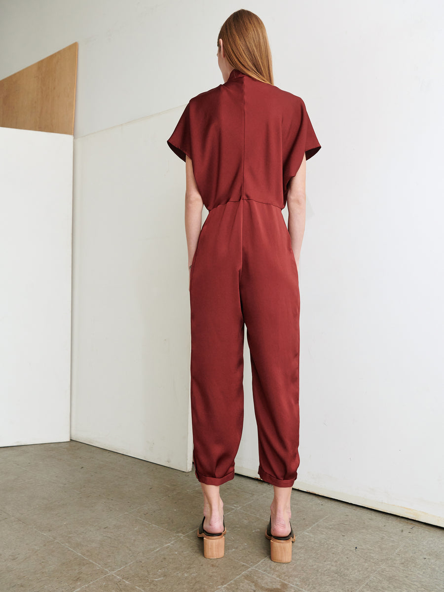 A person with long hair, seen from the back, is wearing the Elia Jumpsuit by Zero + Maria Cornejo, a red short-sleeved outfit crafted from 100% recycled fabric. They pair it with black high-heeled sandals and stand in an open, minimalist indoor space with white walls and a concrete floor.