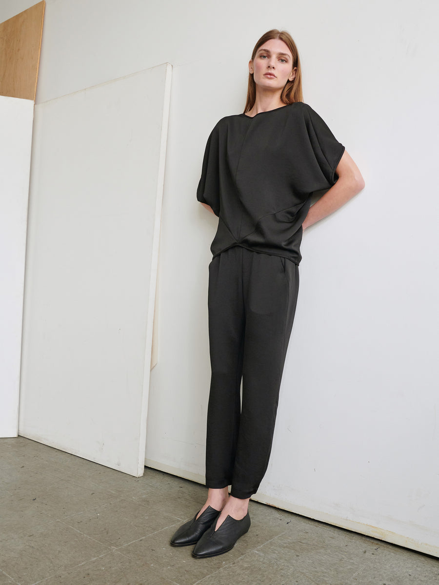 A person with long brown hair stands against a white wall, wearing a loose black top with short sleeves and matching Gabi Trousers from Zero + Maria Cornejo, made of recycled fabric. They have their hands loosely clasped behind their back and are wearing black slip-on shoes. The floor is made of concrete.