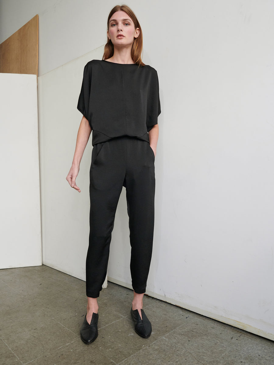 A person with long hair stands against a white wall, wearing a loose-fitting black top and Gabi Trousers from Zero + Maria Cornejo. The trousers feature an elastic waist, and the individual has one hand in their pocket. They are also wearing black shoes, posing in a minimalistic, industrial-style room with a concrete floor.