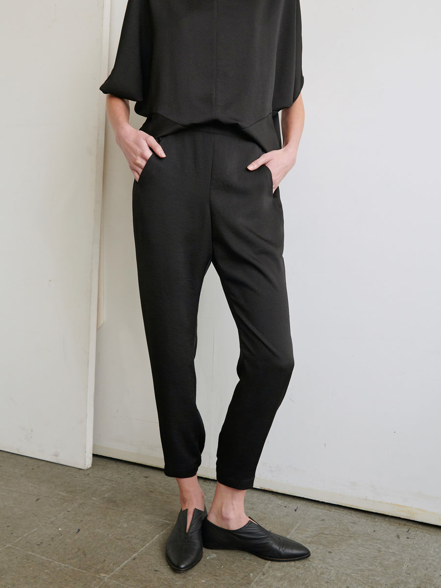 A person is standing against a white wall, wearing a loose black top and Gabi Trousers from Zero + Maria Cornejo made from recycled fabric. Their hands are in the pockets of the pants, which feature an elastic waist. The flooring is a gray, textured material. The person's face is not visible in the image.