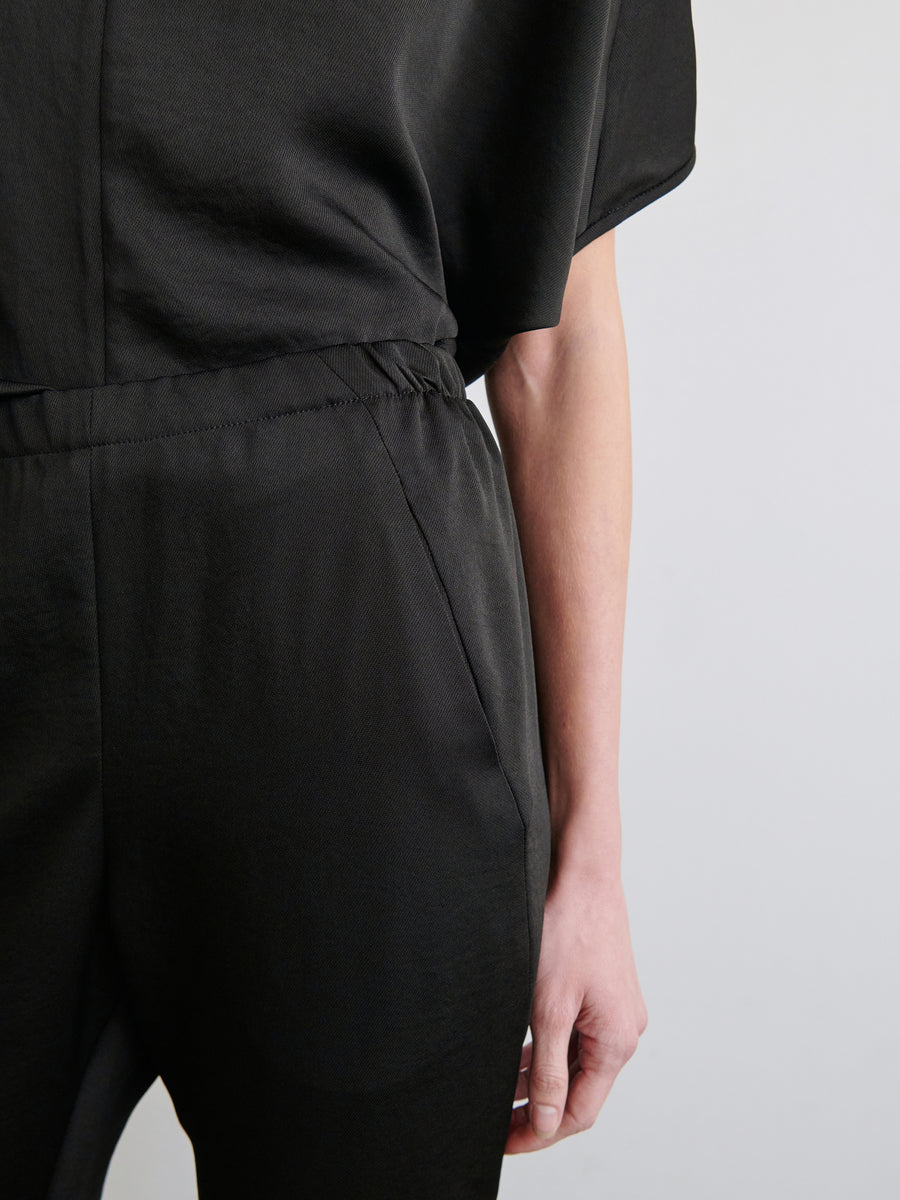 Zoomed in on a person wearing the Gabi Trouser by Zero + Maria Cornejo. The image highlights the torso and upper legs, featuring a cinched elastic waist and smooth, recycled fabric. The person's right arm hangs relaxed at their side, with the hand partially visible.