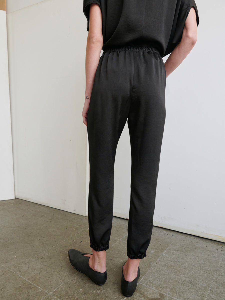 A person is standing against a light-colored wall, facing away from the camera. They are wearing a black short-sleeve top and matching Gabi Trousers from Zero + Maria Cornejo with an elastic waist, along with black slip-on shoes made of recycled fabric. The floor is made of gray concrete.