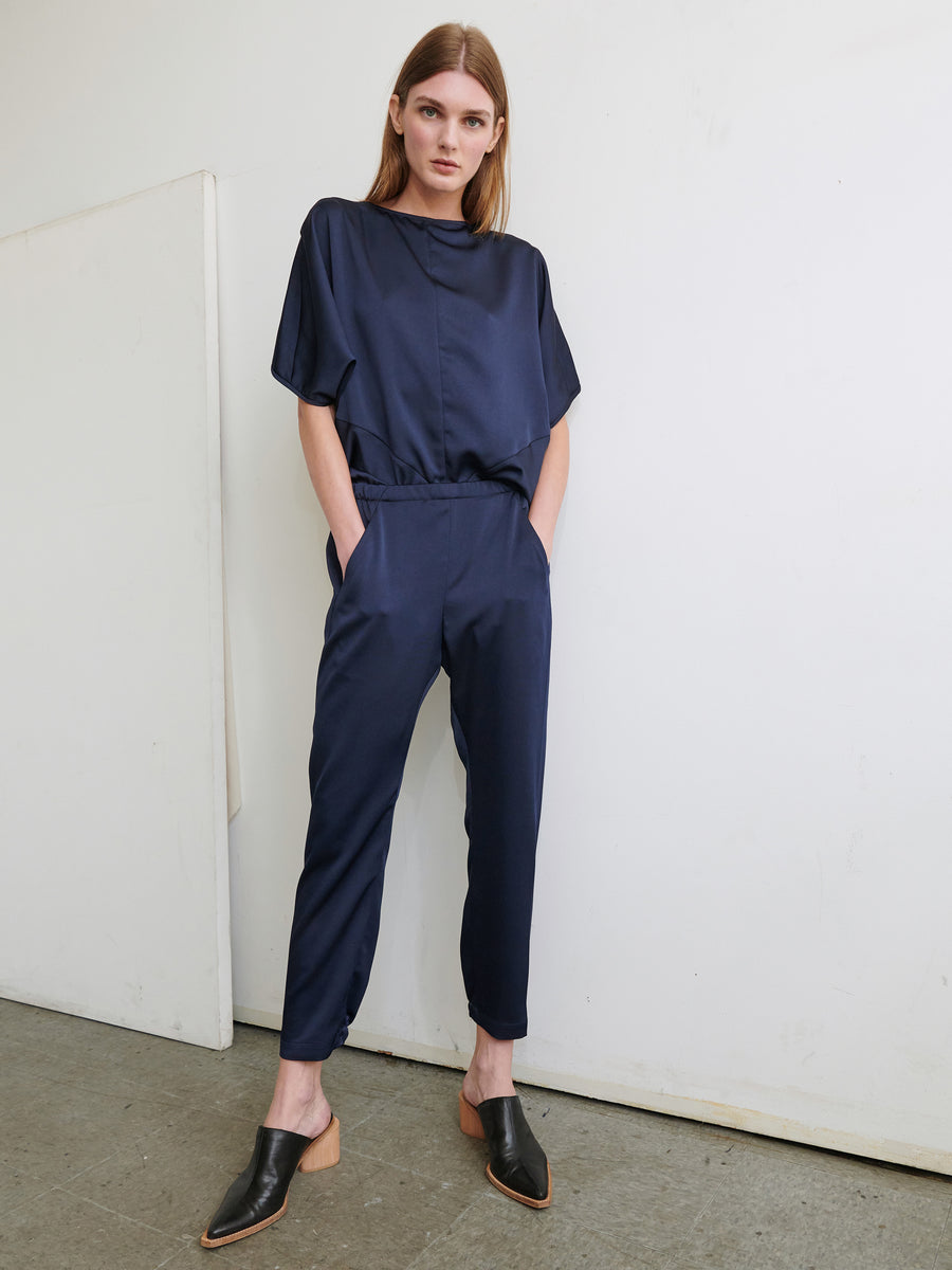 A woman with long brown hair stands in a minimalist room. She is dressed in the Gabi Trouser by Zero + Maria Cornejo, a navy blue satin jumpsuit made from recycled fabric, featuring short sleeves and an elastic waist. Her black slip-on shoes complete the look. Hands in her pockets, she gazes straight ahead.