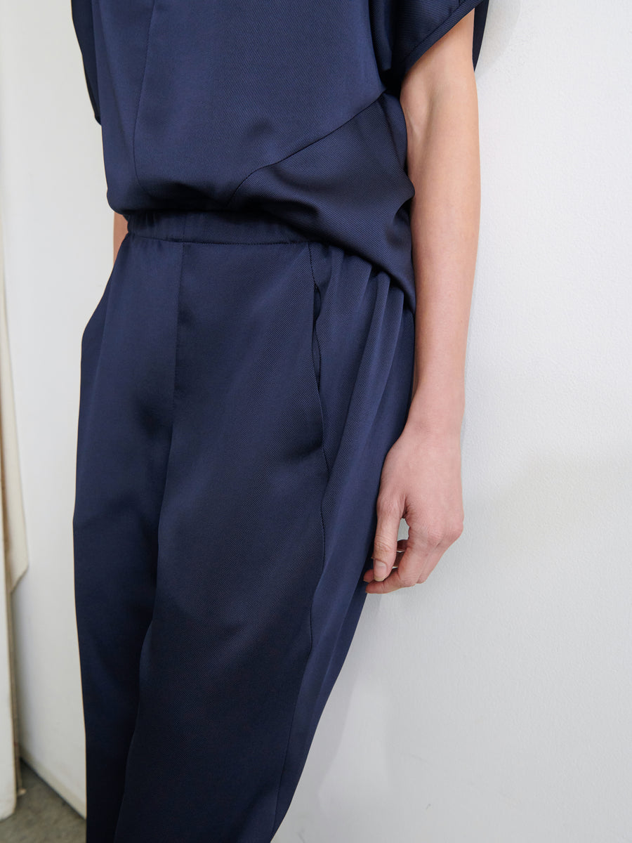 Close-up of a person wearing a navy blue outfit, with their hand relaxed at their side. The outfit consists of a short-sleeved top and high-waisted Gabi Trousers by Zero + Maria Cornejo, made from recycled fabric and featuring hidden pockets. The background is plain white, emphasizing the clothing.