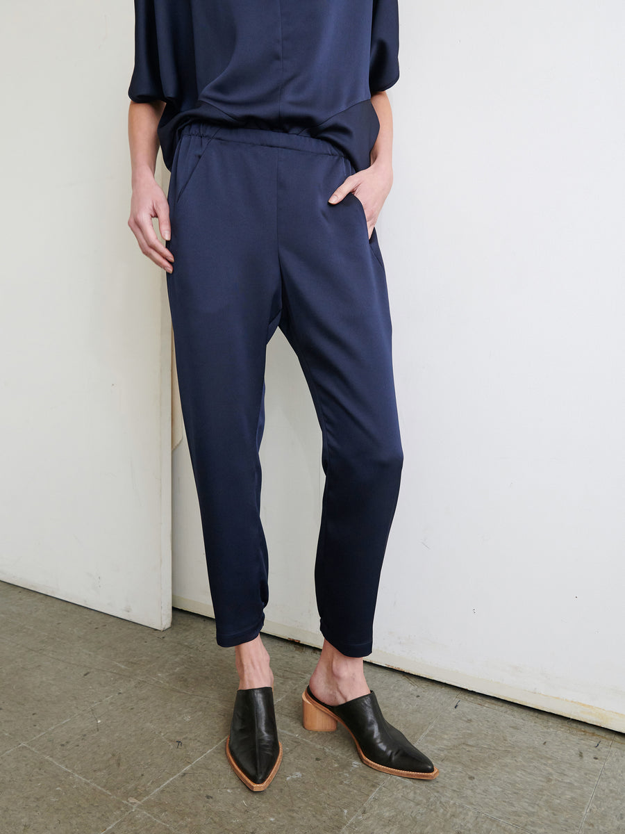 A person dressed in a navy blue ensemble with their hands tucked into the hidden pockets, featuring a loose-fitting top and Gabi Trousers from Zero + Maria Cornejo, crafted from recycled fabric. The individual is also sporting black heeled mules with wooden soles against a plain white wall backdrop.