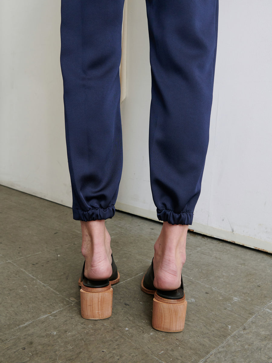 A person stands with their back to the camera, wearing navy blue Gabi Trousers from Zero + Maria Cornejo. These trousers, made from recycled fabric, feature elastic cuffs that expose their ankles and have hidden pockets. They pair the trousers with black open-back shoes that have chunky wooden heels, standing on a gray textured surface against an off-white wall.