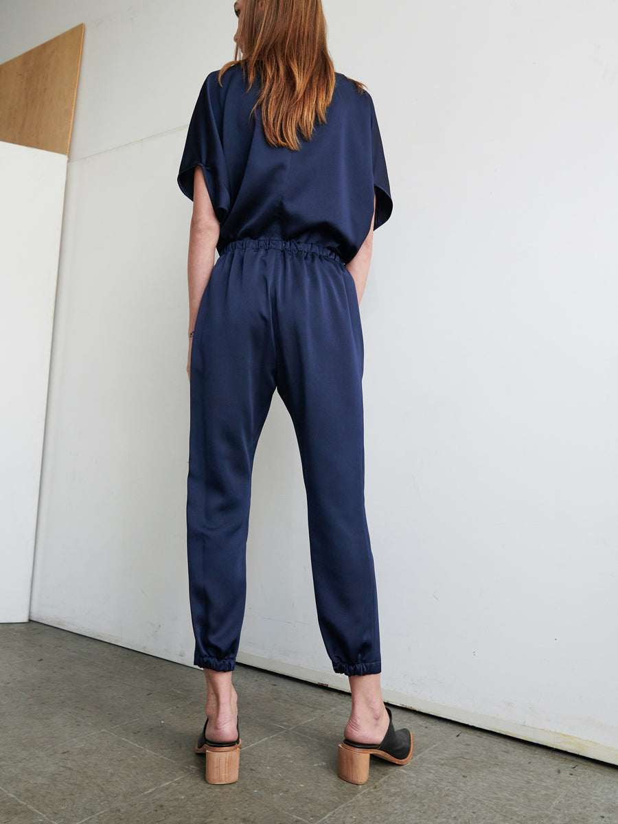 A person with long hair stands facing a wall in a minimalist room, wearing a navy blue short-sleeved top and matching Gabi Trousers by Zero + Maria Cornejo, featuring hidden pockets. Their outfit, paired with black heeled mules, complements the setting's partially visible wooden panel and concrete flooring.