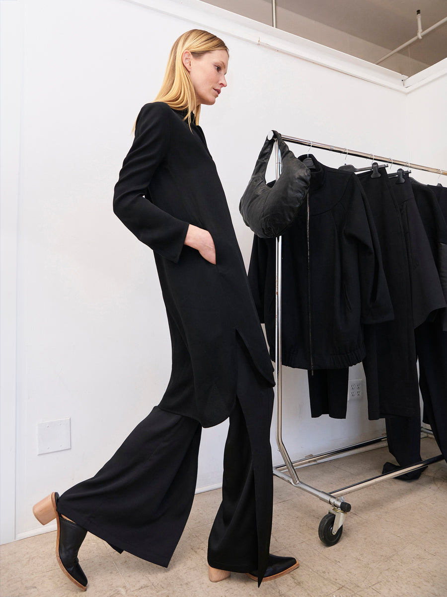 A person with long blonde hair is walking indoors, wearing an all-black outfit made of 100% recycled fabric. The ensemble includes a long coat and the Fin Pant from Zero + Maria Cornejo, featuring wide legs. They are beside a clothes rack displaying black garments, with one hand in their pocket and side slits subtly showing, looking ahead confidently.