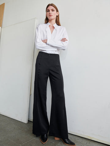 A woman is seen against a plain white wall, dressed in a white blouse with the sleeves folded to her elbows, paired with the high-waisted Fin Pant from Zero + Maria Cornejo, crafted from recycled material and made in New York. She has long, straight hair and gazes confidently at the camera with her arms crossed. Slightly visible are her black shoes.