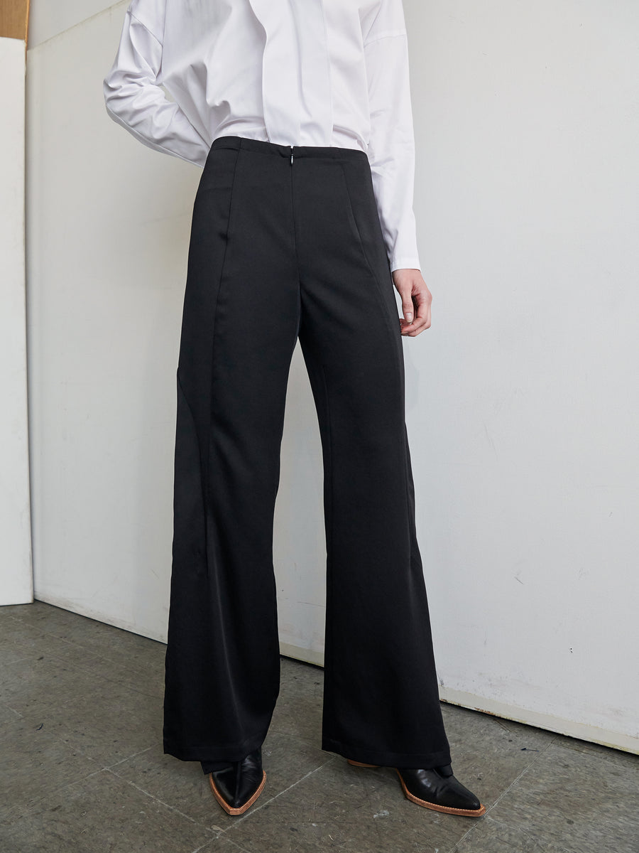 A person is standing indoors wearing a white long-sleeve shirt paired with high-waisted black Fin Pants by Zero + Maria Cornejo. The photo is cropped above the shoulders and below the feet, emphasizing the outfit, which was made in New York. The plain background accentuates the focus on the clothing crafted from 100% recycled fabric.