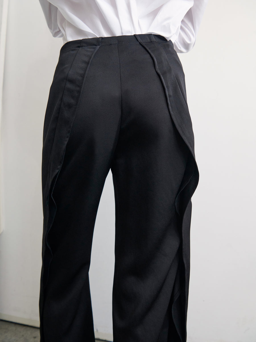 A person is shown from behind wearing the Fin Pant by Zero + Maria Cornejo, black high-waisted trousers featuring a distinctive draped design on the sides, made from 100% recycled fabric. They also sport a white shirt tucked neatly into the trousers. The setting is a simple, white indoor background.