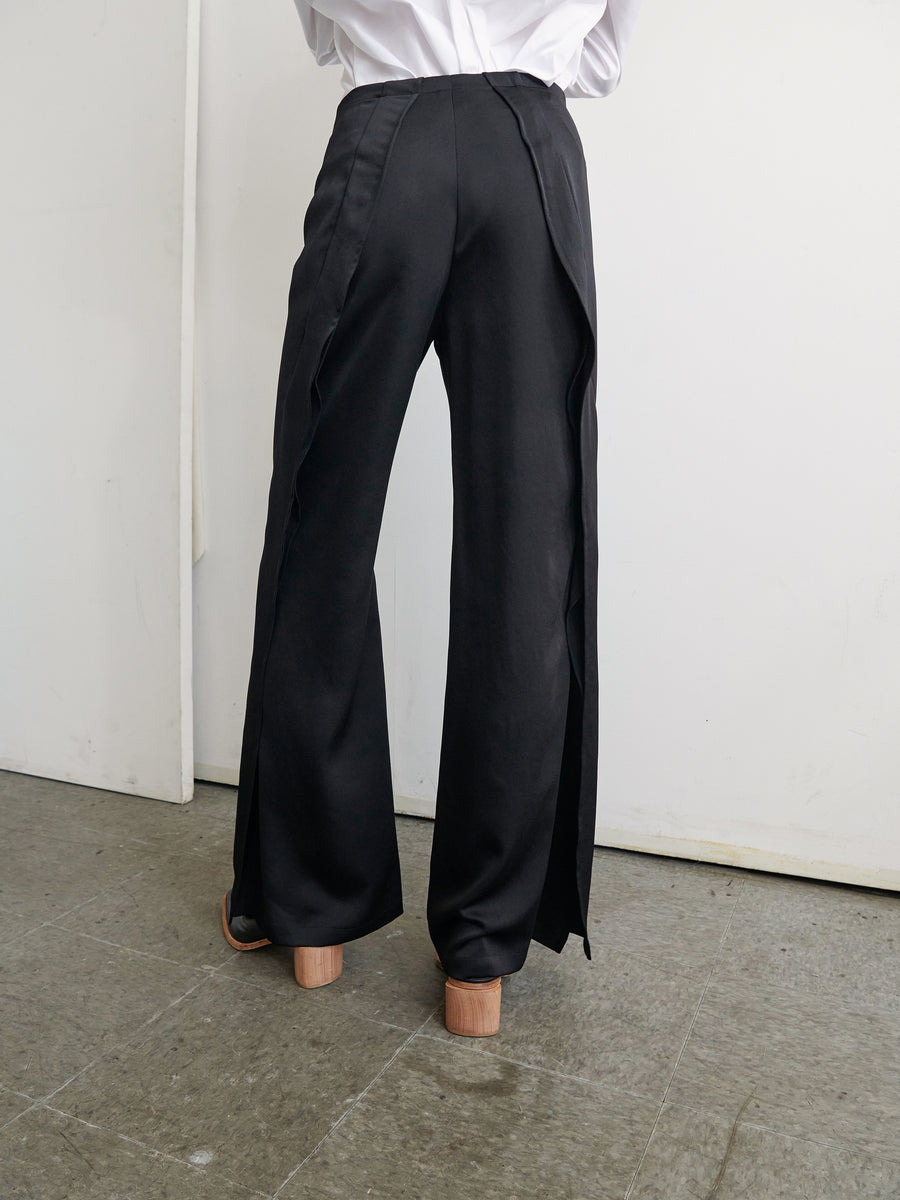 A person in a white shirt is pictured from the waist down, wearing black wide-leg Fin Pants by Zero + Maria Cornejo. The pants feature unique side slits and are made from recycled materials. The floor is gray, and the background is white, highlighting the minimalist and stylish design of this outfit made in New York.