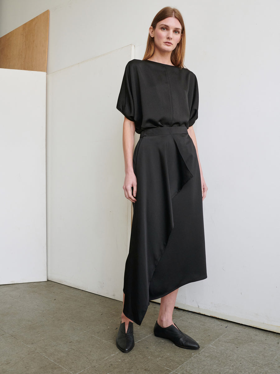 A person with long hair stands against a white wall, wearing a loose-fitting black top with half sleeves and Zero + Maria Cornejo's One Skirt made from recycled fabric. They have black pointed shoes. The background includes a white panel and part of a wooden structure.