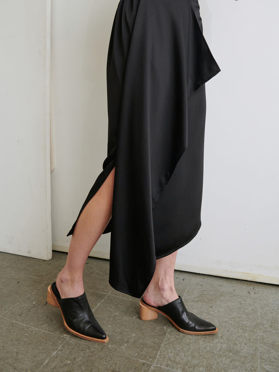 A person is wearing the One Skirt by Zero + Maria Cornejo, a black asymmetrical skirt featuring a high slit and an elastic waist that reveals their left leg. They are paired with black mules boasting wooden heels, standing on a tiled floor against a plain white wall.