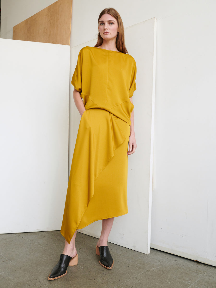 A woman stands against a minimalist background wearing a mustard yellow three-quarter sleeve top and an asymmetrical midi skirt with an elastic waist from Zero + Maria Cornejo's One Skirt collection. She has long, straight hair and is dressed in black, backless mule shoes. The outfit has a draped, elegant design.