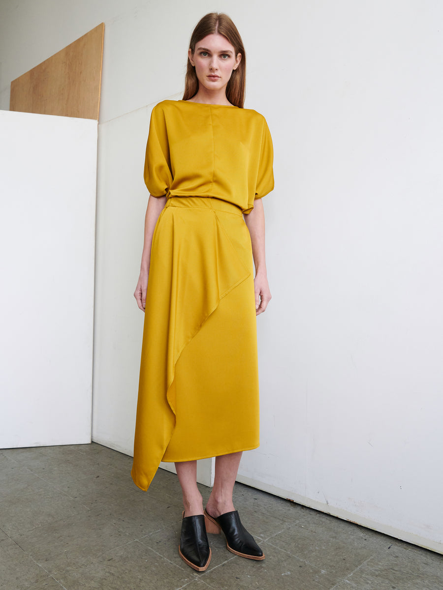 A woman with long brown hair stands against a white wall, wearing the Zero + Maria Cornejo's mustard yellow 'One Skirt' dress. The outfit features an elastic banded waist and short puff sleeves, with an asymmetrical skirt crafted from recycled fabric that boasts an elegant front ruffle. She completes the look with black pointed flats on a concrete floor.