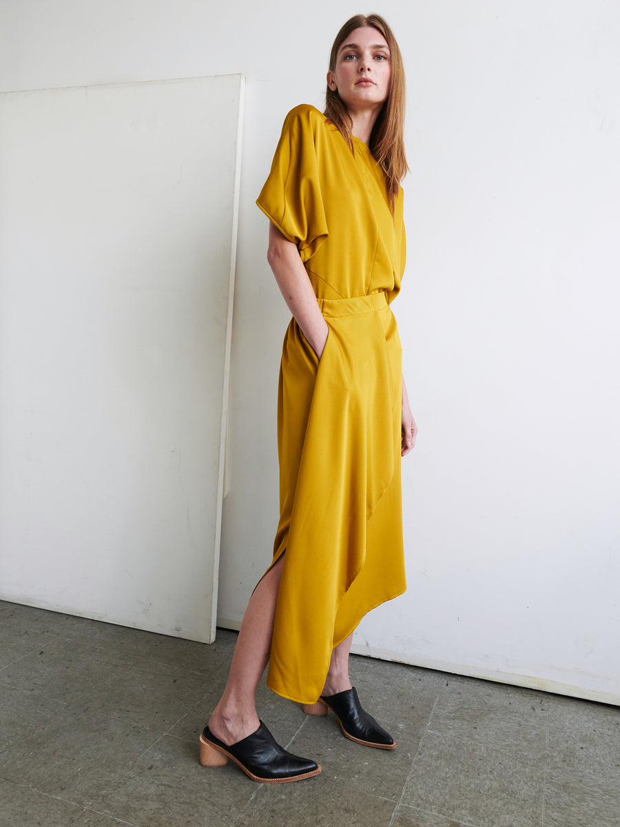 A person with long hair stands against a white wall wearing the One Skirt by Zero + Maria Cornejo, which features an elastic banded waist, short loose sleeves, and an asymmetrical design in mustard yellow. They have their hands in their pockets and are wearing black shoes with a wooden heel. The floor is a grey tone.