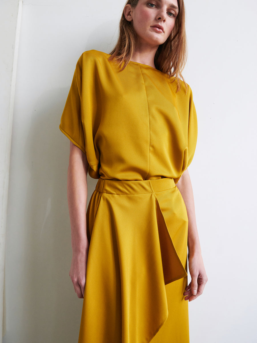 A person with long hair stands against a white wall, wearing a mustard yellow outfit from Zero + Maria Cornejo. The top has loose, cap sleeves, and the One Skirt features an asymmetrical front ruffle and a draped overlay made from recycled fabric. With an elastic banded waist enhancing comfort, their expression is calm and their gaze directed slightly downward.