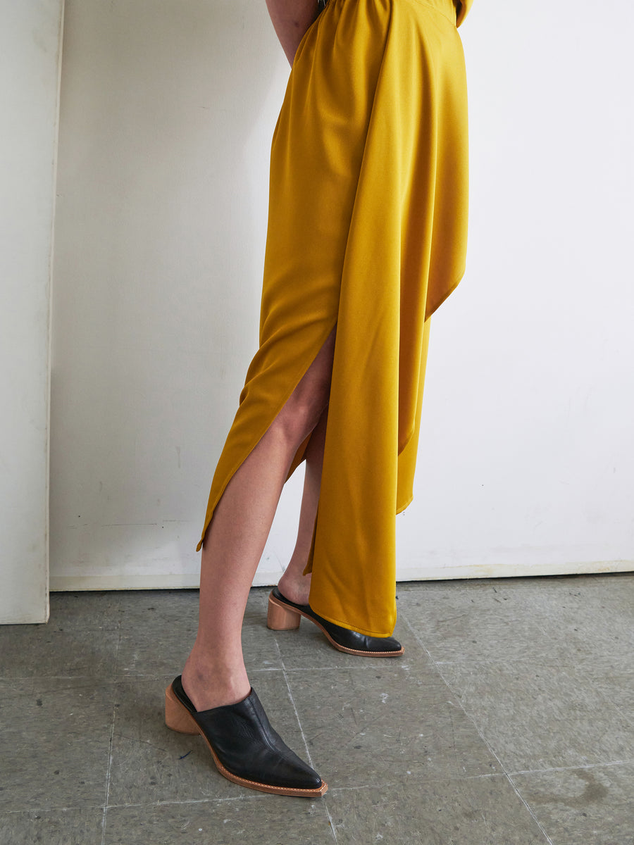 A person stands against a white wall on a gray tiled floor, showcasing the One Skirt by Zero + Maria Cornejo. The mustard-colored skirt features an elastic banded waist and a high slit. They are also wearing black mules with wooden heels. Only the lower half of the body is visible in the image.