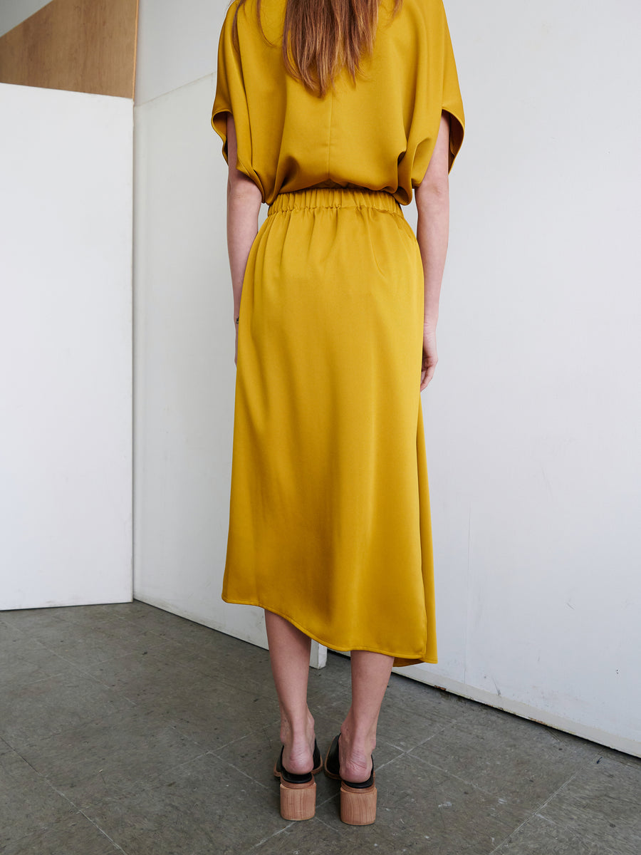 A person with long hair stands with their back to the camera, wearing the One Skirt from Zero + Maria Cornejo, a mustard yellow piece featuring an elastic banded waist, paired with brown heeled sandals. The skirt reaches below their knees, and the scene appears to be indoors with a minimalistic, light-colored background.