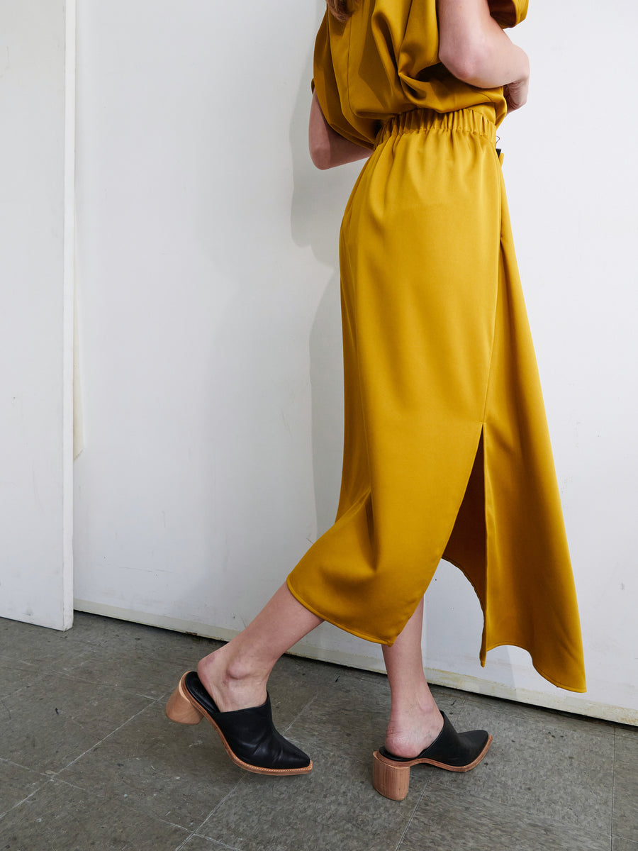 A person is half visible, walking away from the camera. They are wearing the One Skirt by Zero + Maria Cornejo, which is in mustard yellow with an asymmetrical front ruffle and a slit on the side, paired with black open-back shoes featuring wooden block heels. The background is a simple white wall and gray flooring.