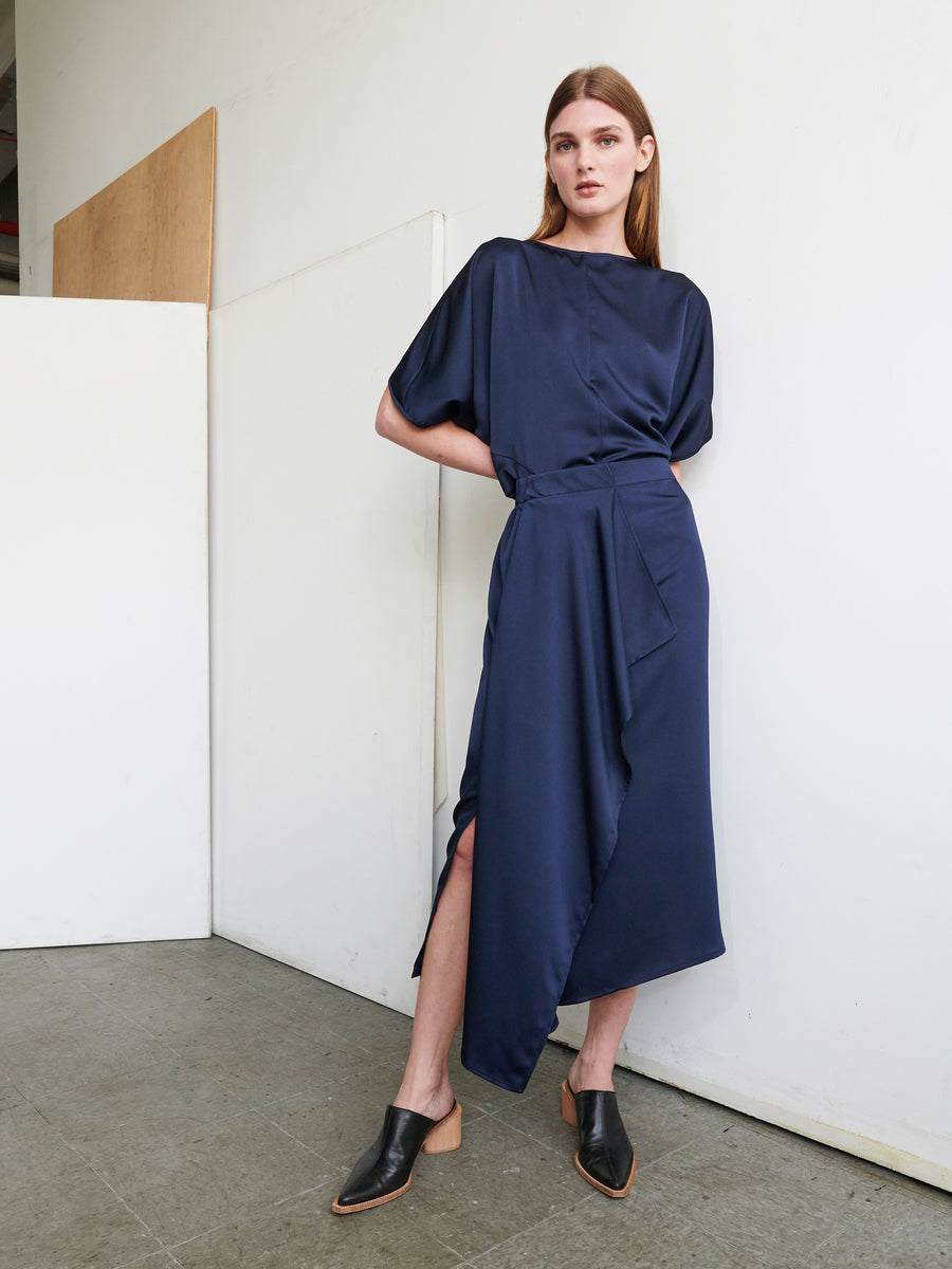 A woman stands against a white wall and a wooden panel, wearing the One Skirt by Zero + Maria Cornejo. The dark blue, asymmetrical dress features loose short sleeves and an elastic banded waist. Its draped skirt, crafted from recycled fabric, complements her shoulder-length brown hair. She completes her look with black slip-on shoes with a low wooden heel and maintains a neutral expression.